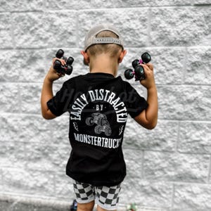 May include: A young child wearing a black t-shirt with the text "Easily Distracted by Monstertrucks" in white. The child is holding toy monster trucks in their hands and wearing a grey baseball cap.