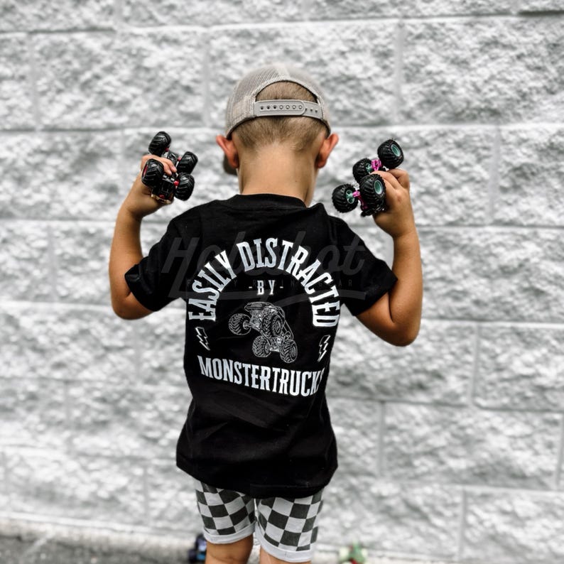 May include: A young child wearing a black t-shirt with the text "Easily Distracted by Monstertrucks" in white. The child is holding toy monster trucks in their hands and wearing a grey baseball cap.
