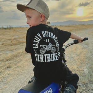 May include: A young boy wearing a black t-shirt with white text that reads "Easily Distracted by Dirtbikes" is riding a blue and black dirt bike.