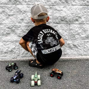 May include: A young boy wearing a black t-shirt with the text "Easily Distracted - By Monstertrucks" in white. He is wearing a grey mesh baseball cap and is sitting on the ground with four toy monster trucks.