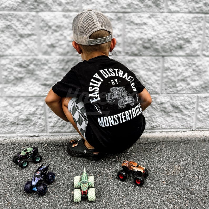 May include: A young boy wearing a black t-shirt with the text "Easily Distracted - By Monstertrucks" in white. He is wearing a grey mesh baseball cap and is sitting on the ground with four toy monster trucks.