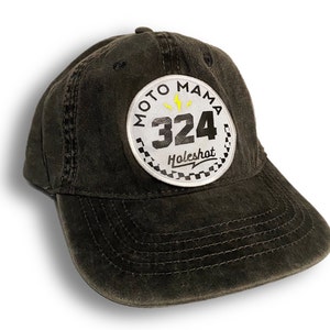 Könnte beinhalten: Eine schwarze Baseballkappe mit einem weißen Patch, auf dem "Moto Mama 324 Holeshot" in schwarzer Schrift mit einem gelben Blitz steht.
