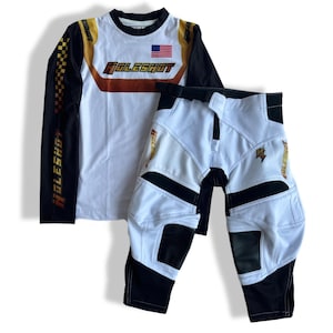 May include: White and black motocross gear set with long-sleeved jersey and pants. The jersey has a gold and orange "Holeshot" logo and an American flag. The pants have a black belt and a gold "Holeshot" logo on the leg.