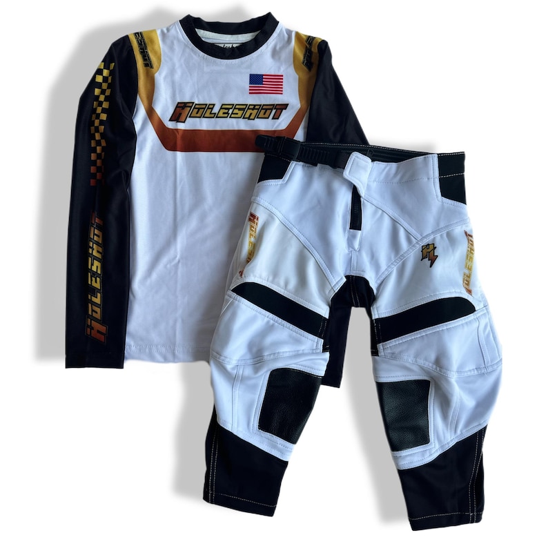 May include: White and black motocross gear set with long-sleeved jersey and pants. The jersey has a gold and orange "Holeshot" logo and an American flag. The pants have a black belt and a gold "Holeshot" logo on the leg.