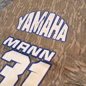 May include: A short-sleeve t-shirt featuring a brown and tan tree bark camouflage pattern. The shirt displays the text YAMAHA in white with a blue outline, followed by the name MANN and the number 31 in large white lettering with blue outlines.