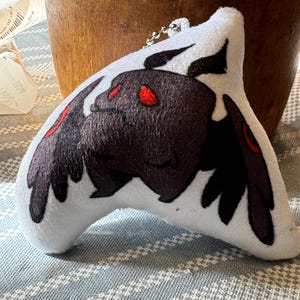 May include: A white plush toy in the shape of a stylized moth. The moth has dark brown wings and body with red eyes and accents. A silver chain is attached to the top of the toy.