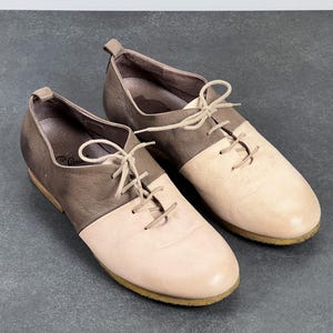 May include: A pair of brown and light pink leather lace-up shoes with a rounded toe. The shoes have a contrasting color design, with the upper part of the shoe being brown and the lower part being light pink.