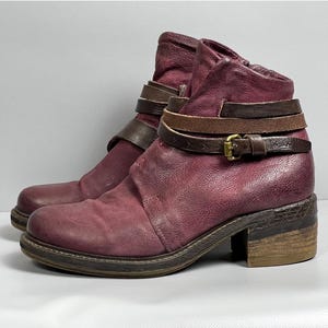 May include: Burgundy leather ankle boots with brown leather straps and a buckle. The boots have a chunky heel and a rounded toe.