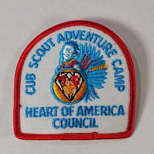 May include: White embroidered patch with red trim. It says "Club Scout Adventure Camp Heart of America Council" with a blue and yellow logo in the center.