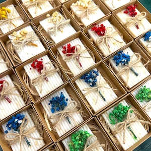 Può includere: Una collezione di piccoli saponi rettangolari, ognuno adornato con un mazzolino di fiori secchi colorati e legato con spago, presentati in scatole individuali di cartone. I saponi sono bianchi con una superficie strutturata.