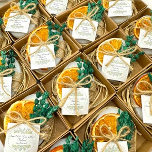 Puede incluir: Arreglo de jabones artesanales en pequeñas cajas de cartón. Cada jabón está decorado con una rodaja de naranja seca, ramitas verdes y una etiqueta atada con hilo. La etiqueta dice: "From Nicole's Shower to Yours! Thank you! 29 June 2023."