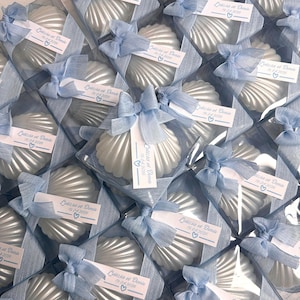 May include: A collection of white shell-shaped items packaged in clear square boxes. Each box is tied with a light blue ribbon and features a white tag printed with the text Letizia et Denis 19.04.2026 and a small blue heart icon.