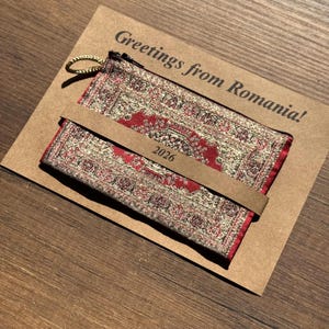 Puede incluir: Un pequeño monedero rectangular y un marcapáginas a juego con un diseño de alfombra oriental roja y dorada. El monedero tiene una cremallera y un anillo dorados. El marcapáginas tiene el año "2026" impreso. Los artículos están en una tarjeta marrón con "Greetings from Romania!"