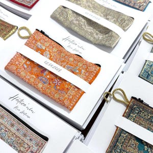 May include: A collection of rectangular zippered pouches featuring intricate woven patterns in orange, gold, blue, and teal. Each pouch has a gold cord loop and is attached to a white card printed with the text Hatice'nin Kina Hatirasi and 02.04.2026.