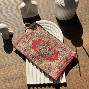 Puede incluir: Un estuche rectangular con estampado rojo y dorado con cierre de cremallera y un lazo dorado. El estuche presenta un diseño detallado de alfombra oriental. El estuche descansa sobre una superficie blanca y curvada.