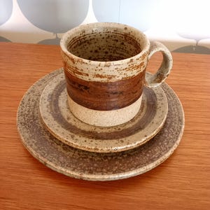 Könnte beinhalten: Ein Keramik-Kaffeebecher- und Untertassen-Set. Der Becher hat ein braunes Band in der Mitte und eine gesprenkelte, cremefarbene Oberfläche. Das Set enthält zwei Teller mit einem ähnlichen gesprenkelten Design und braunen konzentrischen Kreisen. Das Set steht auf einer Holzoberfläche.