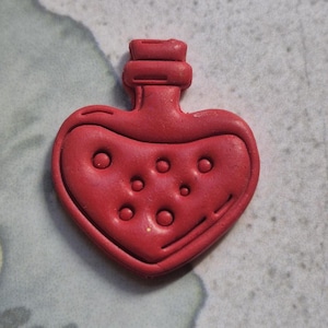 May include: A red, heart-shaped bottle with a cork top. The bottle has a series of circular indentations on the front. The design is a solid, vibrant red colour.