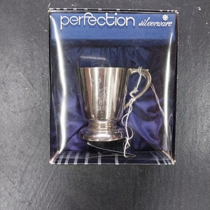 May include: A silver cup with a handle, presented in a box. The box is labelled "perfection silverware". The cup features a pedestal base and an engraved design. The background is a dark blue fabric. The cup is likely made of silver.