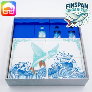 Può includere: Una scatola bianca con interno blu, contenente diverse piastrelle quadrate bianche con un disegno di pesce volante blu e arancione. La scatola presenta anche il testo "FINSPAN ORGANIZER" e un logo di pesce dei cartoni animati.
