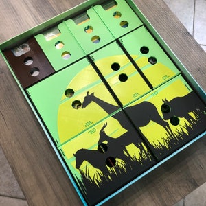 May include: A green and black game box with compartments for storing game pieces. The box features a silhouette of a giraffe, zebra, and antelope against a yellow moon.