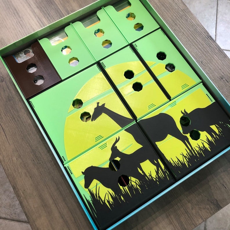 May include: A green and black game box with compartments for storing game pieces. The box features a silhouette of a giraffe, zebra, and antelope against a yellow moon.