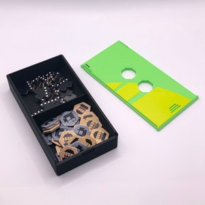 May include: A black box with two compartments. One compartment contains black tile pieces and the other compartment contains gold and silver tokens with a building icon. A green and yellow game board is next to the box.