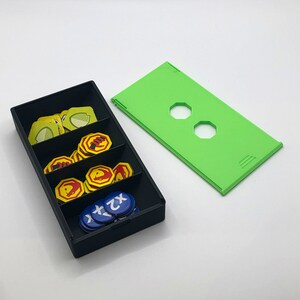 May include: A black storage box with a green lid. The box is filled with small, round, colorful tokens. The tokens are arranged in rows and columns. The box is sitting on a white surface.