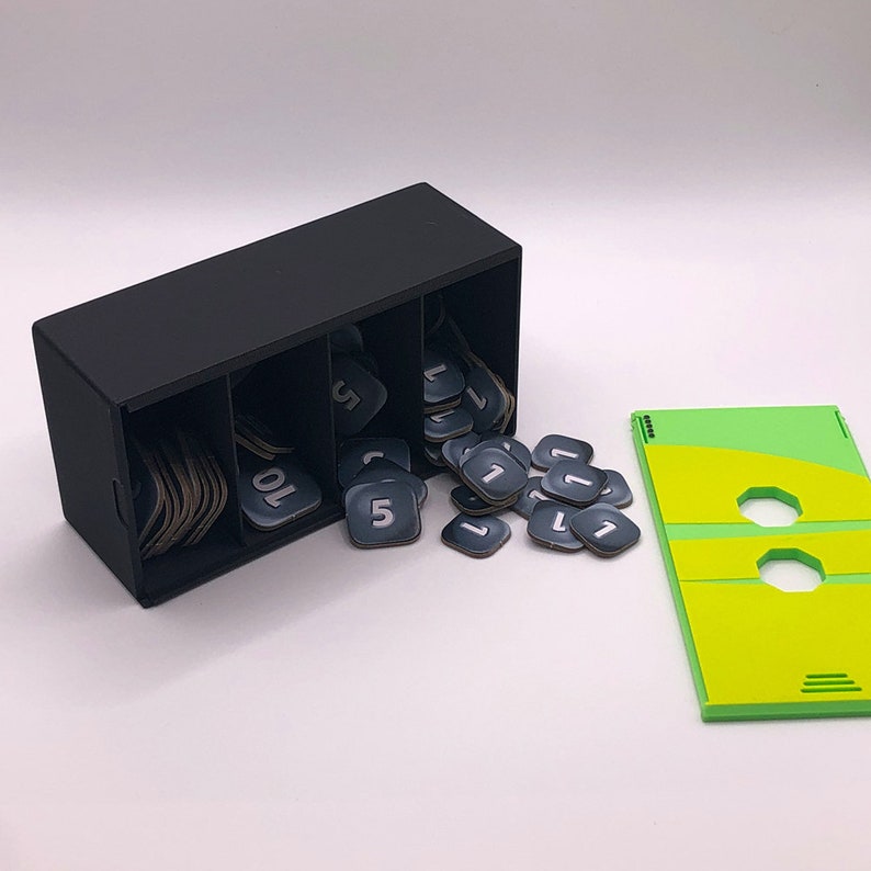May include: A black plastic box with three compartments, filled with small black square tiles with white numbers on them. The tiles are spilling out of the box. There is a green and yellow game board in the background.