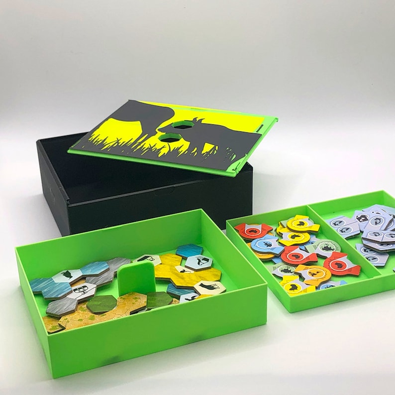 May include: A green and black game box with a lid featuring a silhouette of two animals. The lid is open and the box is filled with small green and yellow tiles. The box is sitting on a white surface.