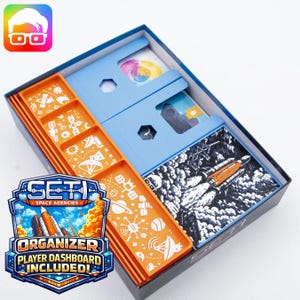 May include: Open board game box with orange and blue components. The box contains cards, player dashboards, and a black and white illustration of a rocket. The game is titled "SETI Space Agencies" and includes an organizer and player dashboard.