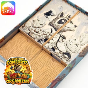 May include: A board game organizer for "Creature Comforts" featuring illustrated animal characters. The box contains wooden-textured game pieces and illustrated tiles with a raccoon, beaver, and rabbit. The organizer has a colorful sticker with the game's title and characters.