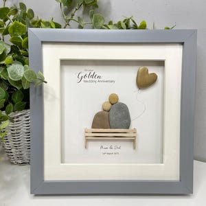 May include: A framed pebble art piece with a couple sitting on a bench, a heart-shaped balloon, and the text "On your Golden Wedding Anniversary Mom & Dad 14th March 1975".