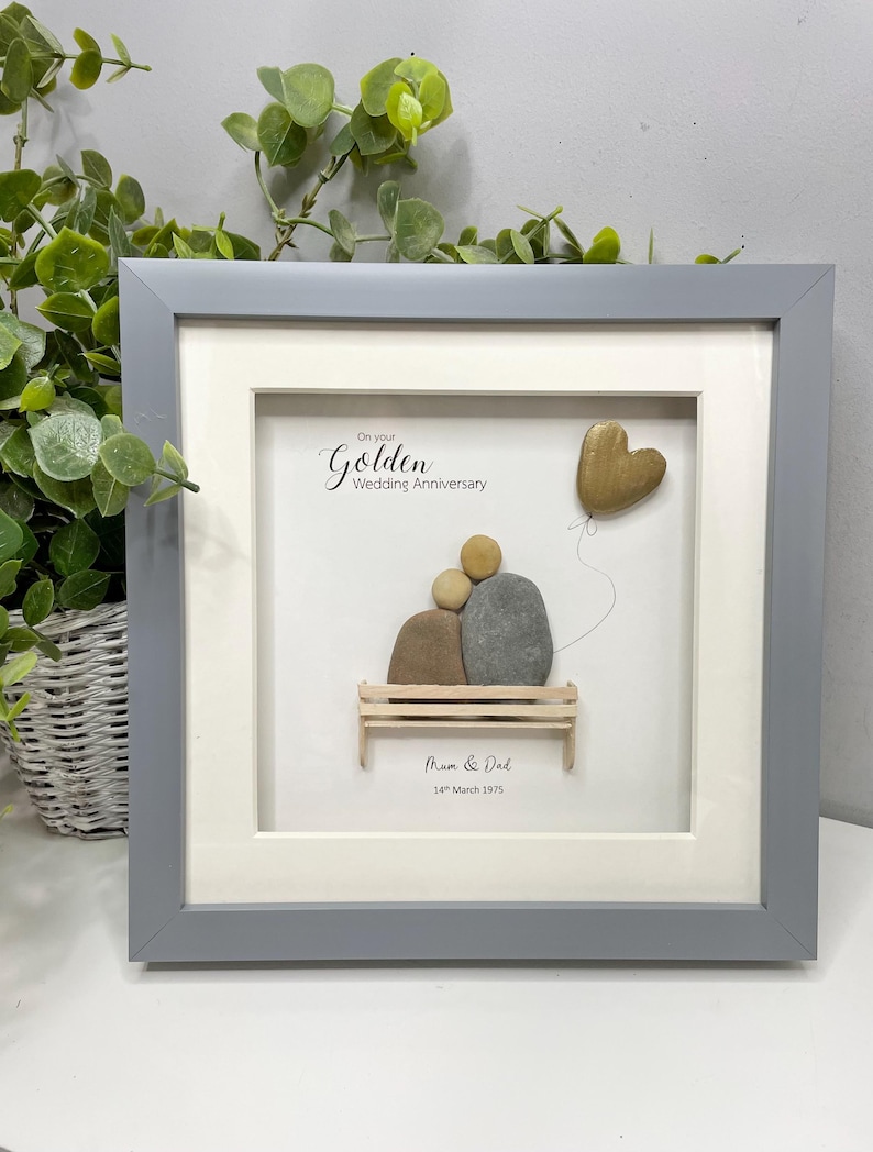 May include: A framed pebble art piece with a couple sitting on a bench, a heart-shaped balloon, and the text "On your Golden Wedding Anniversary Mom & Dad 14th March 1975".