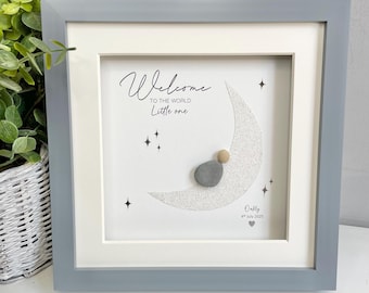 May include: A square gray framed wall art piece featuring a white background with a silver glitter crescent moon. A small gray pebble figure sits on the moon. Text reads: Welcome TO THE WORLD Little one, Oally, 4th July 2025, and a small heart.