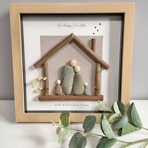 May include: A square light wood frame containing pebble art of a family of four under a twig house structure. The white background features the text "Our Happy Ever After" and the names "Amelie, Ros, Claudie, Sebby" with small heart icons.