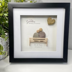 May include: A framed pebble art piece celebrating a golden wedding anniversary. The art depicts a couple sitting on a bench with a golden heart balloon. The text reads "On your Golden Wedding Anniversary" and "Mr & Norman 20th December 1974".