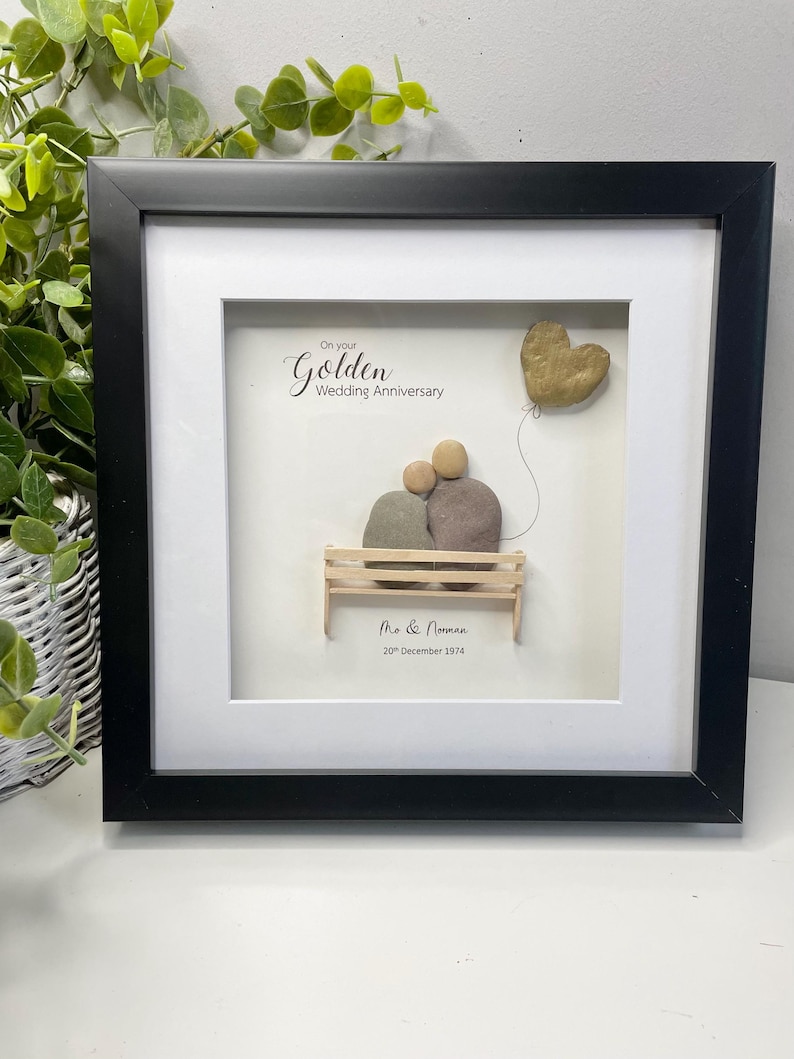 May include: A framed pebble art piece celebrating a golden wedding anniversary. The art depicts a couple sitting on a bench with a golden heart balloon. The text reads "On your Golden Wedding Anniversary" and "Mr & Norman 20th December 1974".