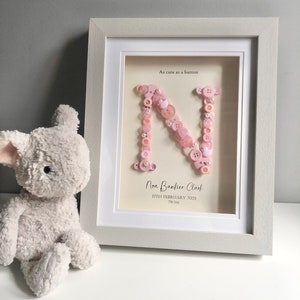 May include: A white frame with a pink button letter N inside. The frame has the text "As cute as a button" and "Nora Bankier Clark 17th February 2023 7lb 5oz".