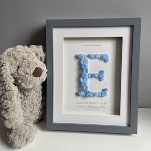May include: A framed wall art piece featuring a large letter E constructed from various shades of blue buttons. The text reads As cute as a button, ELLIOT BENJAMIN AMES, On your Christening Day, Sunday 7th June 2026. A plush beige stuffed animal sits beside it.