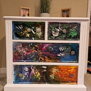 May include: White chest of drawers with four drawers. Each drawer is painted with abstract colourful designs. The dresser has silver hexagonal drawer pulls.