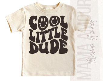May include: A cream colored t-shirt with a black graphic that says "COOL LITTLE DUDE" in a retro font. The graphic also includes two smiley faces.