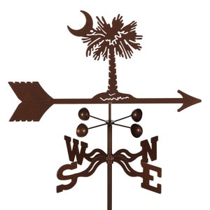 May include: A brown metal weather vane featuring a silhouette of a palmetto tree with a crescent moon, an arrow, and compass directions. The weather vane is mounted on a metal pole.