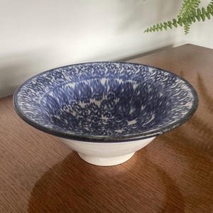 May include: A ceramic bowl with a white base and a wide, shallow blue interior. The blue interior has a speckled pattern. The rim is dark blue. The bowl is sitting on a wooden surface.