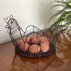 May include: A dark brown wire basket shaped like a chicken with a coiled base. The basket features a wire frame forming the head, neck, and tail of the bird. Inside the wire structure, there are several brown eggs resting on the bottom surface.