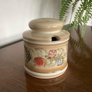 May include: A ceramic jar with a lid, decorated with hand-painted fruits and leaves. The jar is beige and brown, with a white band at the base. The lid has a small opening for a spoon. The jar is suitable for storing preserves.