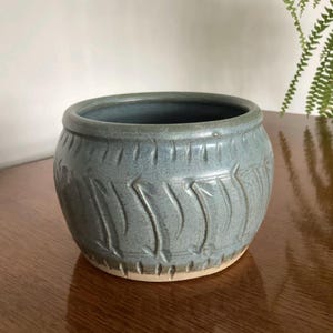 May include: A blue-grey ceramic plant pot with a carved, textured pattern. The round pot has a wide opening and a light-coloured base. The exterior features vertical, curved lines. Ideal for small plants or succulents.