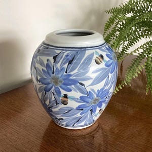 May include: A ceramic vase with a rounded body and a wide opening, featuring a hand-painted pattern of blue flowers, green leaves, and small bees with black and yellow stripes against a white background, finished with a smooth light grey rim.