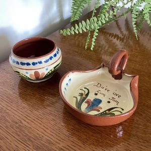 May include: A small round ceramic bowl with blue and green floral patterns and a matching shallow dish with a handle, featuring the text Du'ee ave Zum Jam painted in brown script along with a blue and green floral design on the interior.