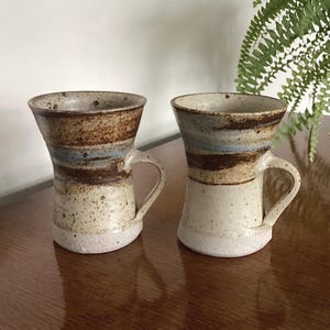 May include: A pair of handcrafted ceramic mugs with a distinctive flared shape. The mugs have a speckled, off-white base, with bands of brown, blue, and tan. Each mug has a curved handle and a rustic, artisanal appearance.
