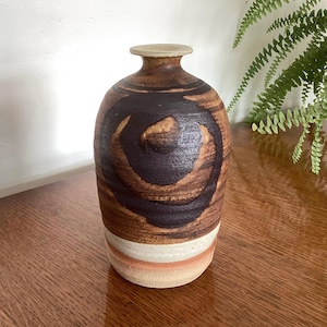 May include: A cylindrical ceramic vase with a narrow neck and a flared rim. The body features a dark brown spiral pattern over a textured brown surface, transitioning to a smooth white band and a light orange base with a matte, earthy finish.